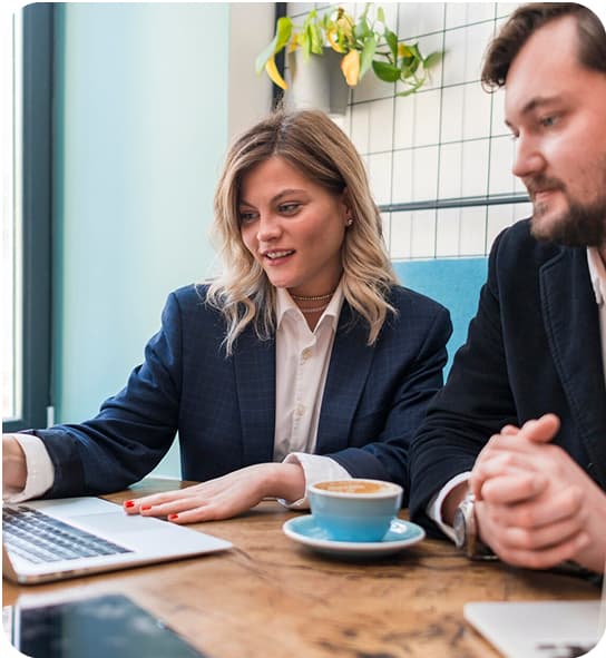 A woman and a man discuss work at a table with a laptop and coffee